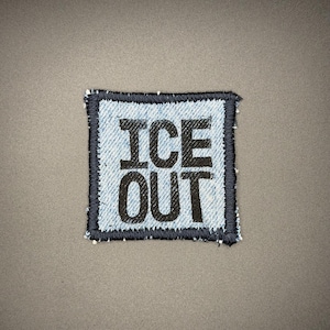 May include: Square denim patch with the words "ICE OUT" in bold black letters. The patch has a dark blue border and is set against a gray background. The patch is likely for clothing or accessories.