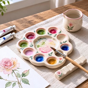 Peut inclure: Une palette de peinture en céramique en forme de fleur avec dix puits circulaires contenant diverses couleurs d'aquarelle. La palette présente de petits motifs floraux peints, un pichet d'eau assorti, un pinceau en bois et une peinture de rose.