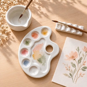 May include: A white ceramic watercolor palette with six circular wells containing traces of pink, green, blue, and yellow paint, a matching white water bowl with a brush, and a white ceramic brush rest holding a wooden paintbrush.