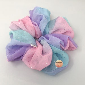 May include: A pastel-colored scrunchie with a gradient effect, featuring shades of pink, blue, and purple. The fabric is adorned with small, silver glitter dots. The scrunchie is a hair accessory.