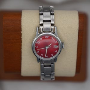 Könnte beinhalten: Silberfarbene Timex-Uhr mit rotem Zifferblatt und Datumsfenster. Die Uhr hat ein silbernes Armband und wird auf einer beigen Oberfläche präsentiert. Der Durchmesser des Zifferblatts beträgt ca. 2,5 cm.