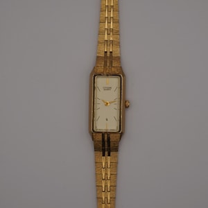 May include: A gold-toned Citizen watch with a rectangular face and a gold link band. The watch face is cream-colored with gold hands and hour markers. The watch has a date display at the bottom of the face.