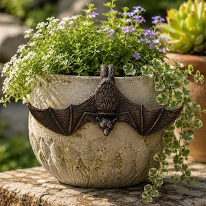 Peut inclure: Un pot de fleurs rond en pierre claire avec une surface texturée et un ornement de chauve-souris sculpté en brun foncé fixé sur le devant. Le pot est rempli d'une variété de petites plantes vertes, de fleurs blanches et de fleurs violettes.