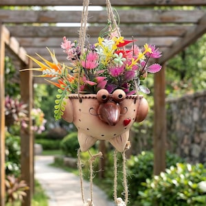 May include: A beige ceramic hanging planter shaped like a bird with large eyes, a pointed beak, and a small red heart detail. The planter is filled with a variety of colourful artificial flowers and foliage, suspended by jute twine ropes.