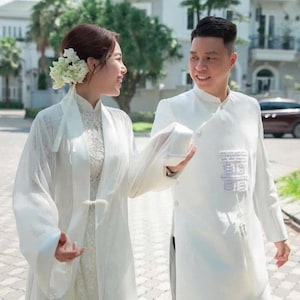 May include: A white traditional long-sleeved tunic with a high collar and a white sheer outer robe featuring a tie closure. The tunic displays a rectangular embroidered patch on the chest. A white floral hair accessory is worn on the side of the head.