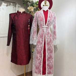 May include: Two traditional Vietnamese outfits on display. The first is a long-sleeved, burgundy tunic with a mandarin collar. The second is a red dress with a white lace overlay and a matching headpiece. Both outfits are displayed on mannequins.