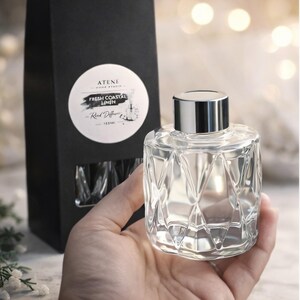 Puede incluir: Un frasco difusor de caña de vidrio transparente con tapa plateada se sostiene en una mano. La botella tiene un diseño geométrico. Una caja negra con una etiqueta blanca que dice "Fresh Coastal Ligne" está en el fondo. El producto es de "ATENE".