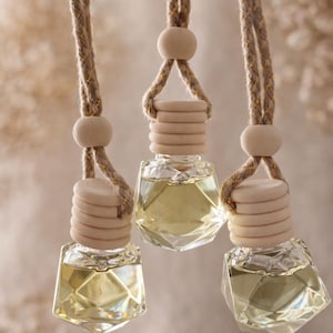 Puede incluir: Tres ambientadores colgantes para coche. Cada uno tiene una botella de vidrio transparente de forma geométrica llena de líquido amarillo. Las botellas están rematadas con una tapa de madera y una cuenta de madera, suspendidas por una cuerda beige.