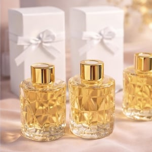 May include: Three glass bottles with gold-colored lids and a geometric pattern, filled with a golden liquid. Two white gift boxes with white ribbon bows are in the background. The bottles are on a pink surface.