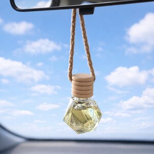 May include: A car air freshener with a geometric glass bottle filled with yellow liquid, hanging from a rearview mirror. The bottle has a wooden cap and is attached to a braided rope. The background shows a blue sky with clouds.