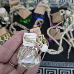 Op de afbeelding: Een heldere, geometrisch gevormde glazen fles met een witte dop en een gevlochten koord. De fles is gevuld met een lichtgekleurde vloeistof en maakt deel uit van een collectie luchtverfrissers met houten accenten en decoratieve koorden.