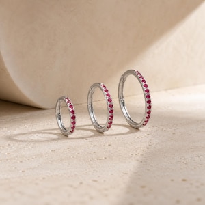 May include: Three silver-toned hoop earrings of varying sizes, each featuring a row of small, round, vibrant red gemstones set along the outer edge. The hoops have a hinged closure mechanism and a polished metallic finish, displayed in a row.