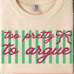 May include: Cream-colored t-shirt with the phrase "too pretty to argue" in pink script, set against a green striped background. A pink bow is on the right side of the text. The shirt is folded on a denim surface.