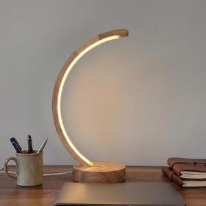 Può includere: Una moderna lampada da scrivania in legno con un design curvo e illuminato. La lampada ha una base circolare e un bagliore caldo. La lampada è su una scrivania in legno con un laptop, una tazza con penne e un taccuino rilegato in pelle.