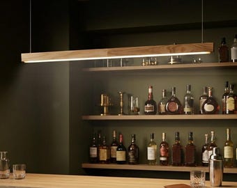 May include: A long, linear wooden pendant light fixture suspended by thin wires above a wooden bar counter. The light features a warm, integrated LED strip that illuminates the bar area, which includes shelves stocked with various glass liquor bottles.