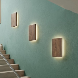May include: Three rectangular wall sconces featuring a dark brown wood grain finish. Each fixture is mounted flush against the wall and emits a soft, warm glow from behind the wooden panel, creating a halo effect on the textured green wall surface.