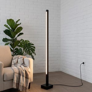 May include: A modern black floor lamp with a rectangular base and a vertical light strip. The lamp is positioned next to a beige armchair with a plaid throw blanket and a large green plant. The lamp is plugged into a wall outlet.