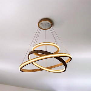 Può includere: Lampadario moderno in legno con tre anelli intersecati. Gli anelli sono in legno scuro e presentano illuminazione a LED integrata. L'apparecchio è sospeso al soffitto tramite sottili fili, creando un design di illuminazione contemporaneo.