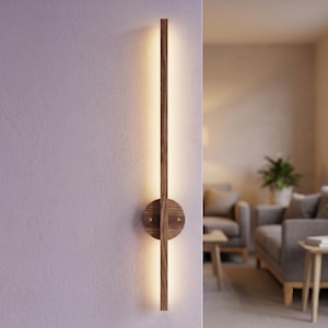 May include: A vertical wall-mounted light fitting featuring a long, narrow wooden bar with a visible grain pattern. A circular wooden base plate is centred on the bar, secured with two small metallic accents. The fitting emits a warm, soft glow.