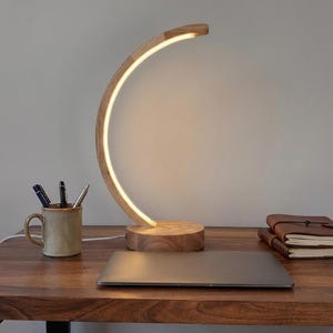 May include: A wooden desk lamp with a curved, illuminated design. The lamp has a circular base and a warm, white light. The lamp is on a wooden desk with a laptop, a mug with pens, and a leather-bound notebook.