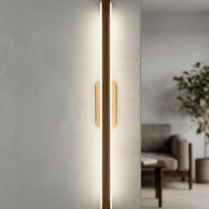 Peut inclure: Une applique murale moderne au design vertical. Elle présente un cadre en bois foncé avec une bande lumineuse centrale. Deux éléments en bois clair sont positionnés en dessous. L'applique est fixée sur un mur texturé de couleur claire.