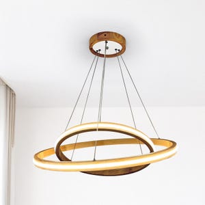 Op de afbeelding: Een moderne houten hanglamp met twee elkaar kruisende, verlichte ringen. De lamp hangt aan het plafond met dunne draden en heeft een cirkelvormige houten basis en kap. Het licht geeft een warme, uitnodigende gloed.