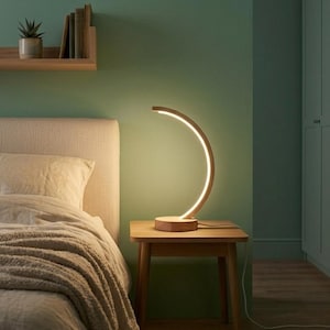 Könnte beinhalten: Eine halbmondförmige Tischleuchte mit einem hellen Holzsockel und Rahmen. Die innere Kurve der Leuchte verfügt über einen integrierten LED-Lichtstreifen, der ein warmes, sanftes Licht abgibt. Die Leuchte steht auf einem hölzernen Nachttisch.