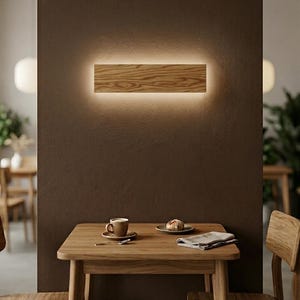 May include: A rectangular wooden wall light fixture with a warm, glowing backlight. The fixture is mounted on a brown wall above a wooden table. A cup of coffee and a pastry are on the table.