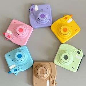 May include: A collection of colorful, square-shaped toy cameras in pink, purple, yellow, green, blue, and tan. Each camera has a circular lens and a small viewfinder. The cameras are arranged in a circular pattern.