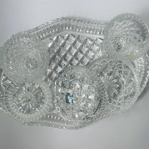 May include: A clear, cut-glass serving set featuring a tray and several small dishes. The tray has a diamond pattern and a scalloped edge. The dishes have various decorative patterns, including starburst and spiral designs. The set is designed for serving appetizers or small portions.
