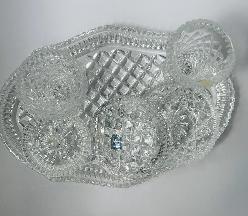 May include: A clear, cut-glass serving set featuring a tray and several small dishes. The tray has a diamond pattern and a scalloped edge. The dishes have various decorative patterns, including starburst and spiral designs. The set is designed for serving appetizers or small portions.