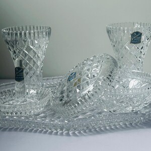May include: A clear crystal serving set featuring two vases, a large oval platter, two small bowls, and a covered dish. The pieces have a diamond-cut pattern and are arranged on a white surface. Each piece has a blue and white sticker.