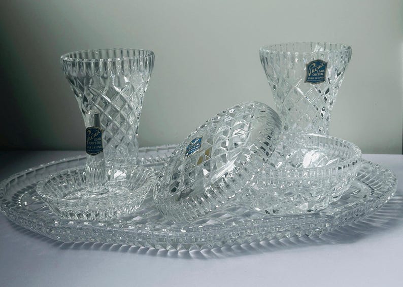 May include: A clear crystal serving set featuring two vases, a large oval platter, two small bowls, and a covered dish. The pieces have a diamond-cut pattern and are arranged on a white surface. Each piece has a blue and white sticker.