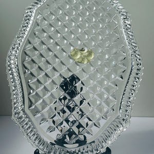 May include: A clear, oval-shaped glass serving tray with a diamond pattern and a decorative, ridged border. The tray is supported by a black stand. A gold sticker is in the center of the tray.