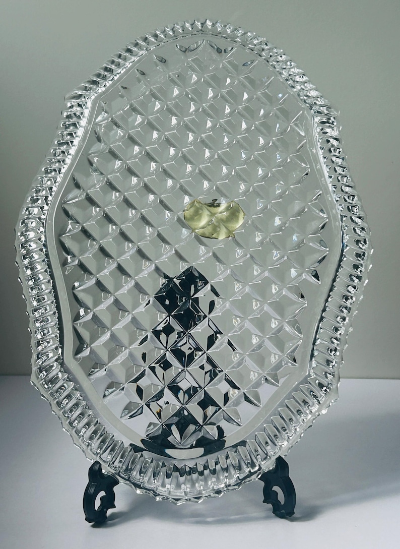 May include: A clear, oval-shaped glass serving tray with a diamond pattern and a decorative, ridged border. The tray is supported by a black stand. A gold sticker is in the center of the tray.
