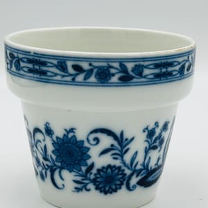 May include: A white ceramic flower pot featuring a blue floral pattern. The design includes a band of repeating flowers and vines around the upper rim, with larger, detailed blue floral motifs and scrolling foliage decorating the main body.