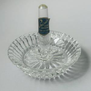 May include: Clear lead crystal ring holder with a round base and a central post. The base has a ribbed design around the edge and a starburst pattern in the center. A blue and gold label reads "Phoenix Lead Crystal Over 24% PbO".