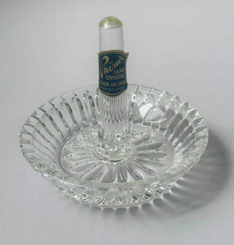 May include: Clear lead crystal ring holder with a round base and a central post. The base has a ribbed design around the edge and a starburst pattern in the center. A blue and gold label reads "Phoenix Lead Crystal Over 24% PbO".