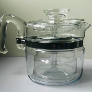 Könnte beinhalten: Eine Kaffeebereiter-Perkolator-Kanne aus klarem Glas mit transparentem Deckel, einem stabilen Glasgriff und einem silberfarbenen Metallband um die Mitte. Im Inneren befindet sich ein Glaskorb-Einsatz, die Glasoberfläche ist glatt und reflektierend.