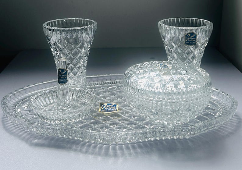 May include: A clear crystal vanity set with diamond-cut patterns. The set includes two vases, a lidded jar, a small dish, and an oval tray. The crystal pieces have a sparkling appearance, reflecting light. The set is ideal for displaying perfumes or jewelry.