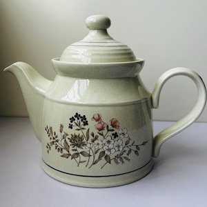 Puede incluir: Una tetera de cerámica de color crema claro con acabado moteado, que presenta una ilustración floral en tonos marrón, rosa y blanco. La tetera incluye una tapa a juego con pomo, un pico curvo y un asa lateral.