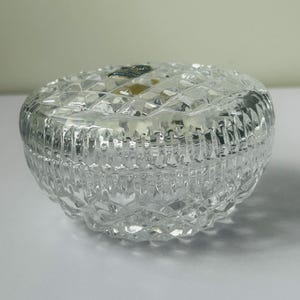 May include: A clear, round crystal trinket box with a diamond-cut pattern. The lid fits snugly on the base, and the box is designed to reflect light. The box is a decorative item.