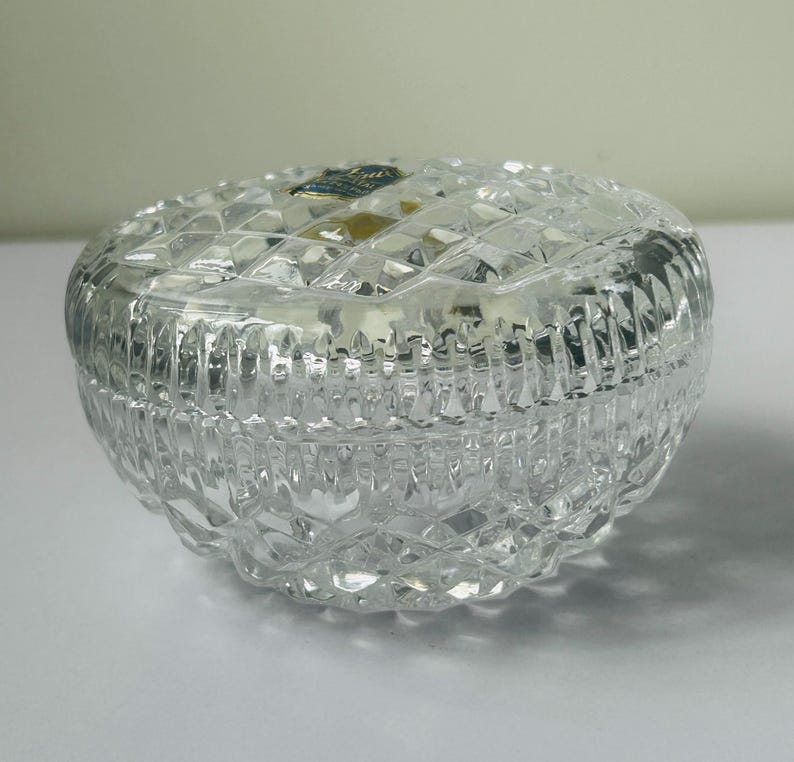 May include: A clear, round crystal trinket box with a diamond-cut pattern. The lid fits snugly on the base, and the box is designed to reflect light. The box is a decorative item.