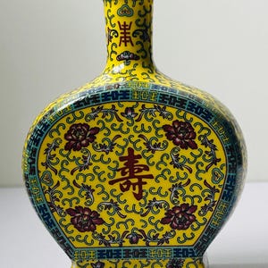 May include: A yellow ceramic moon flask vase featuring intricate floral patterns in dark red, blue, and green. The centre displays a prominent dark red Chinese character, with a decorative geometric border framing the design and a narrow neck.