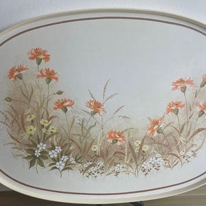 May include: An oval-shaped serving tray with a cream-coloured base and a thin brown border. The centre features a printed illustration of orange cornflowers, small yellow blossoms, and white flowers amidst brown grasses and foliage.