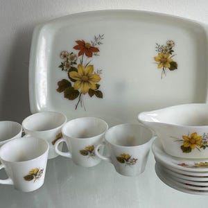 Puede incluir: Un juego de té de vidrio lechoso blanco que incluye una bandeja rectangular, seis tazas, una salsera y seis platillos. Cada pieza está decorada con un patrón floral de flores amarillas y naranjas con hojas verdes y pequeñas flores blancas.