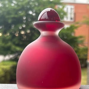 Può includere: Una piccola bottiglia in vetro arrotondata con una finitura satinata rosso intenso. La bottiglia presenta un corpo liscio e bulboso che si restringe verso un collo stretto, sormontato da un tappo sferico rosso scuro lucido e brillante.