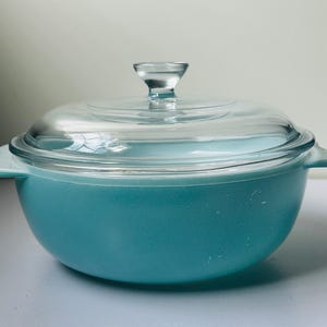 Peut inclure: Une cocotte ronde turquoise avec des poignées latérales intégrées et un couvercle en verre transparent assorti doté d'un bouton central. La base du plat présente une finition opaque et lisse, tandis que le couvercle est entièrement transparent.
