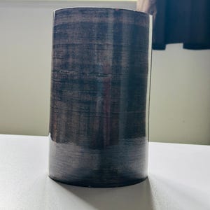 Puede incluir: Un jarrón cilíndrico con un acabado texturizado en gris oscuro y marrón. El jarrón está sobre una superficie blanca, con una sombra proyectada hacia la derecha. El jarrón tiene un aspecto brillante.