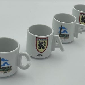 May include: Four white ceramic mugs arranged in a diagonal line. Each mug features a handle shaped like a letter, spelling out CAFE. Two mugs display a crest with the text FLANDRE and the word Lille, while two show a building illustration.